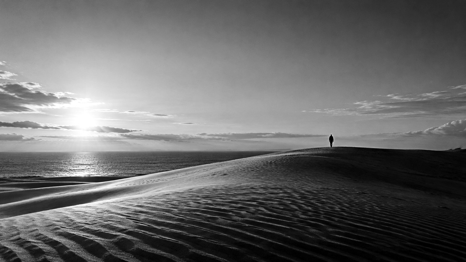 風紋が刻まれた海辺の大きな砂丘と、遠くの稜線を歩く人影（イメージ）